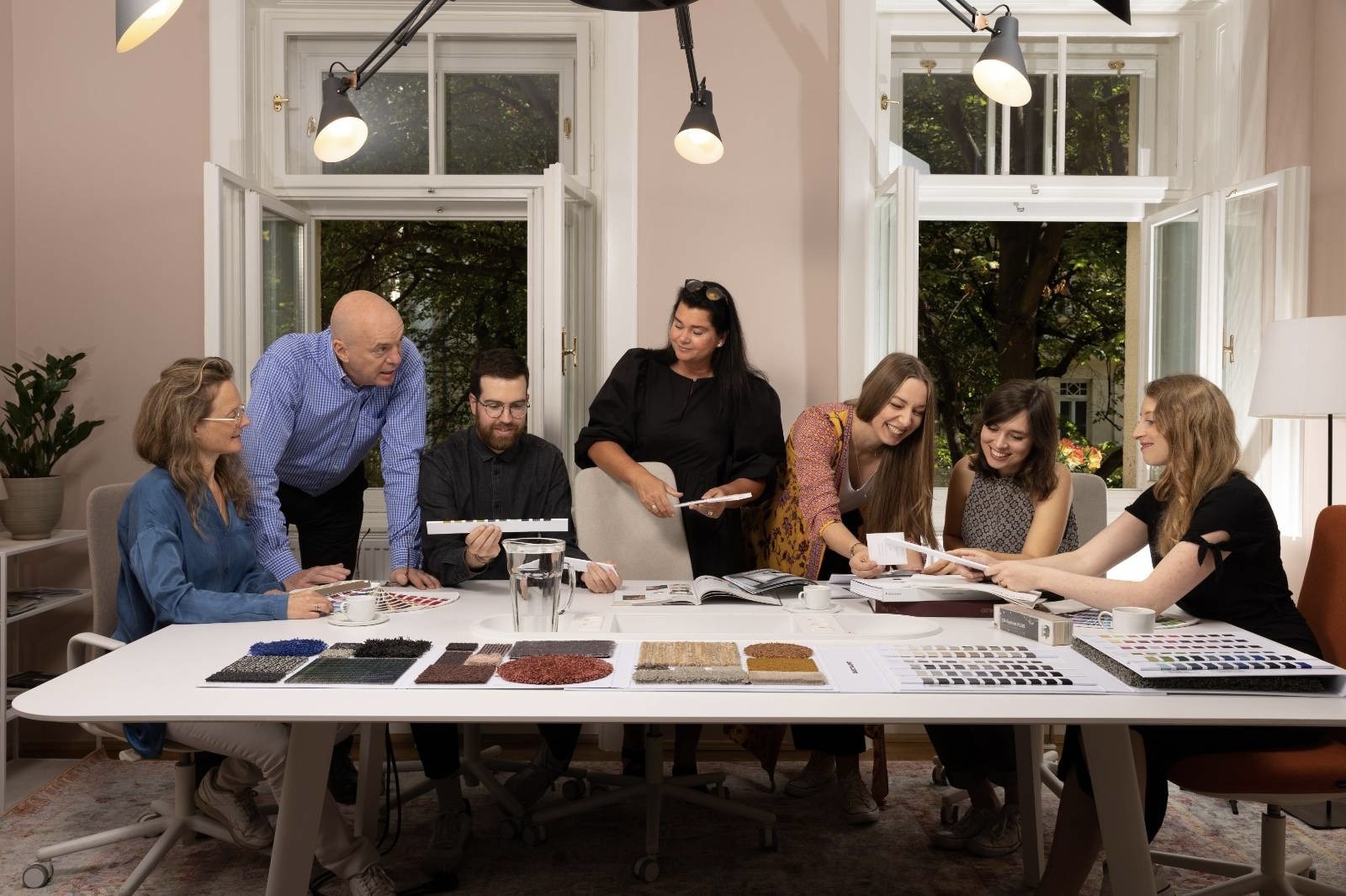 mixed team of women and men sitting in bright room at table filled with fabrics and color samples and open windows