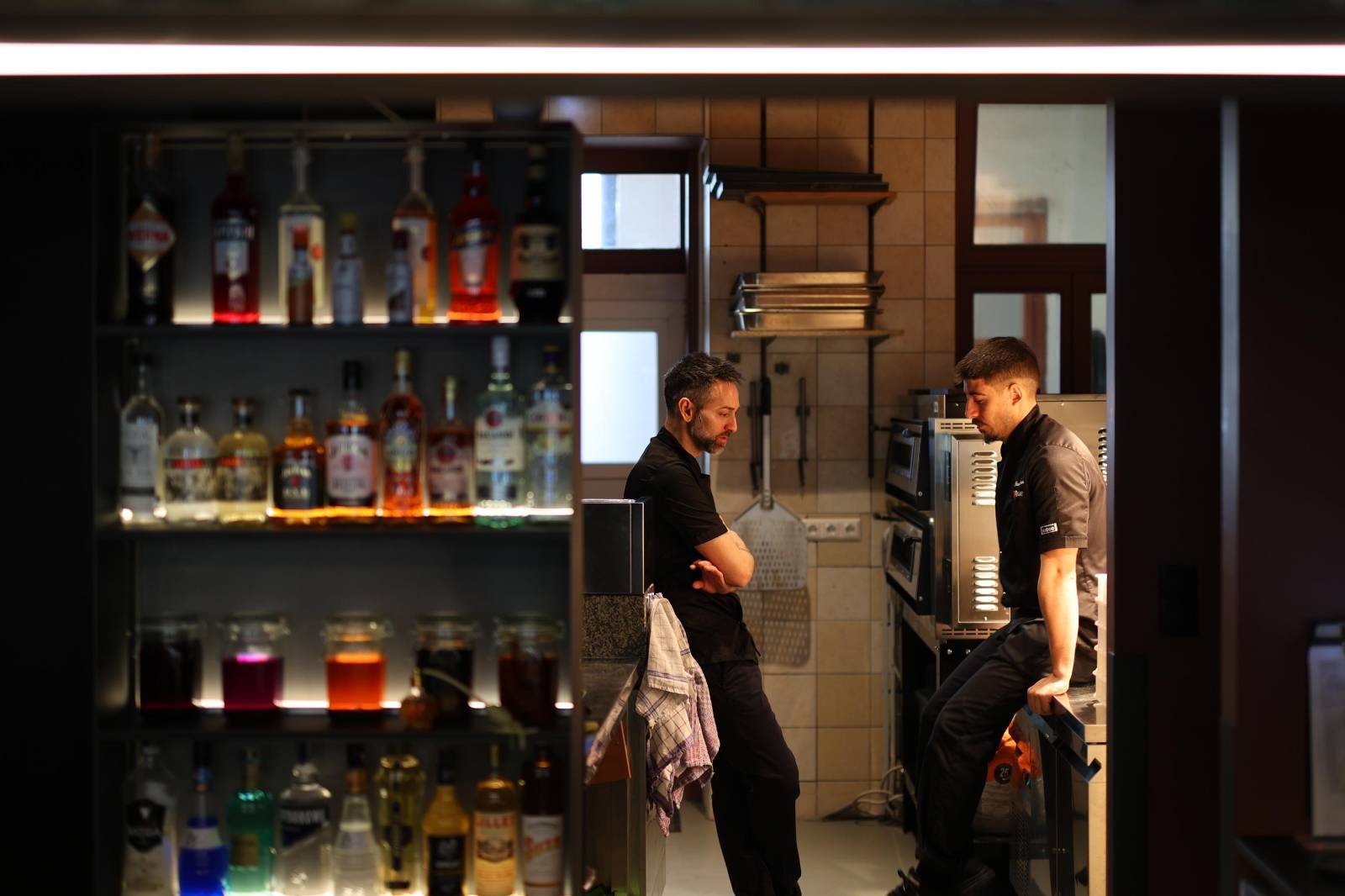 two male chefs standing in kitchen, shelf with spirits in front